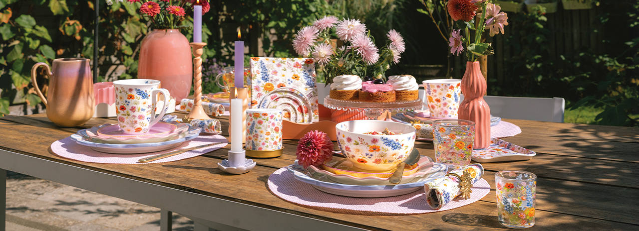 Üppig gedeckter Gartentisch mit 'Blooming summer' Geschirr, bunten Blumen und Kerzen.