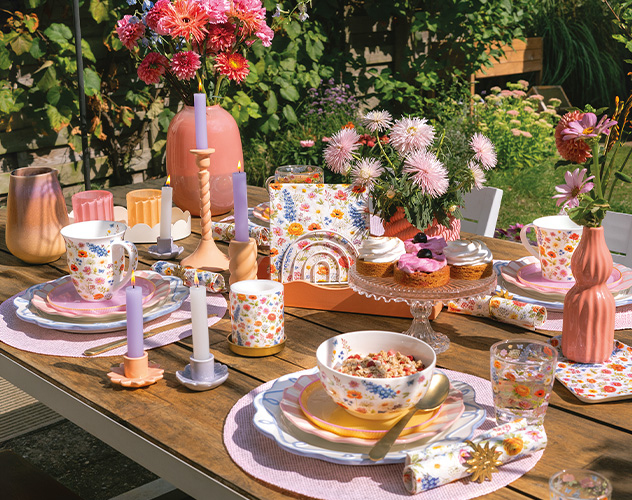 Uitbundig gedekte tuintafel met 'Blooming summer' servies, kleurrijke bloemen en kaarsen.