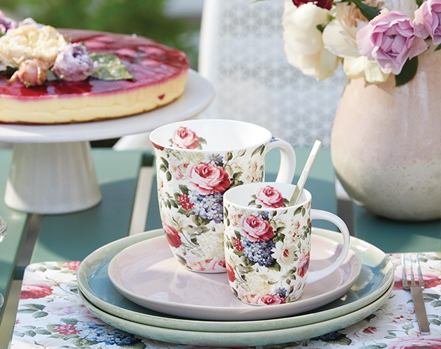 Mokken met 'Rosemary' rozenprint op een stapel borden naast een taart en verse bloemen.