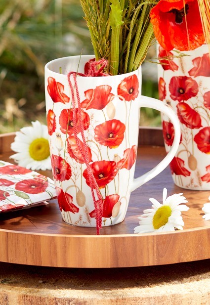 Close-up of a detailed garden picnic setup on grass, featuring a vintage suitcase, teepee, and various items adorned with red poppy patterns from the 'Poppy all over' series.