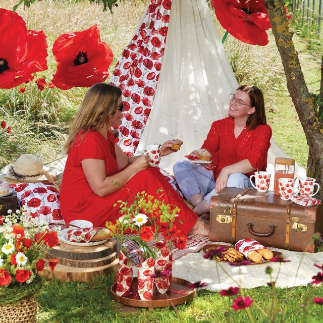 Two women enjoying a garden picnic in the 'Poppy all over' series, with a teepee tent and table setting featuring red poppy patterns.