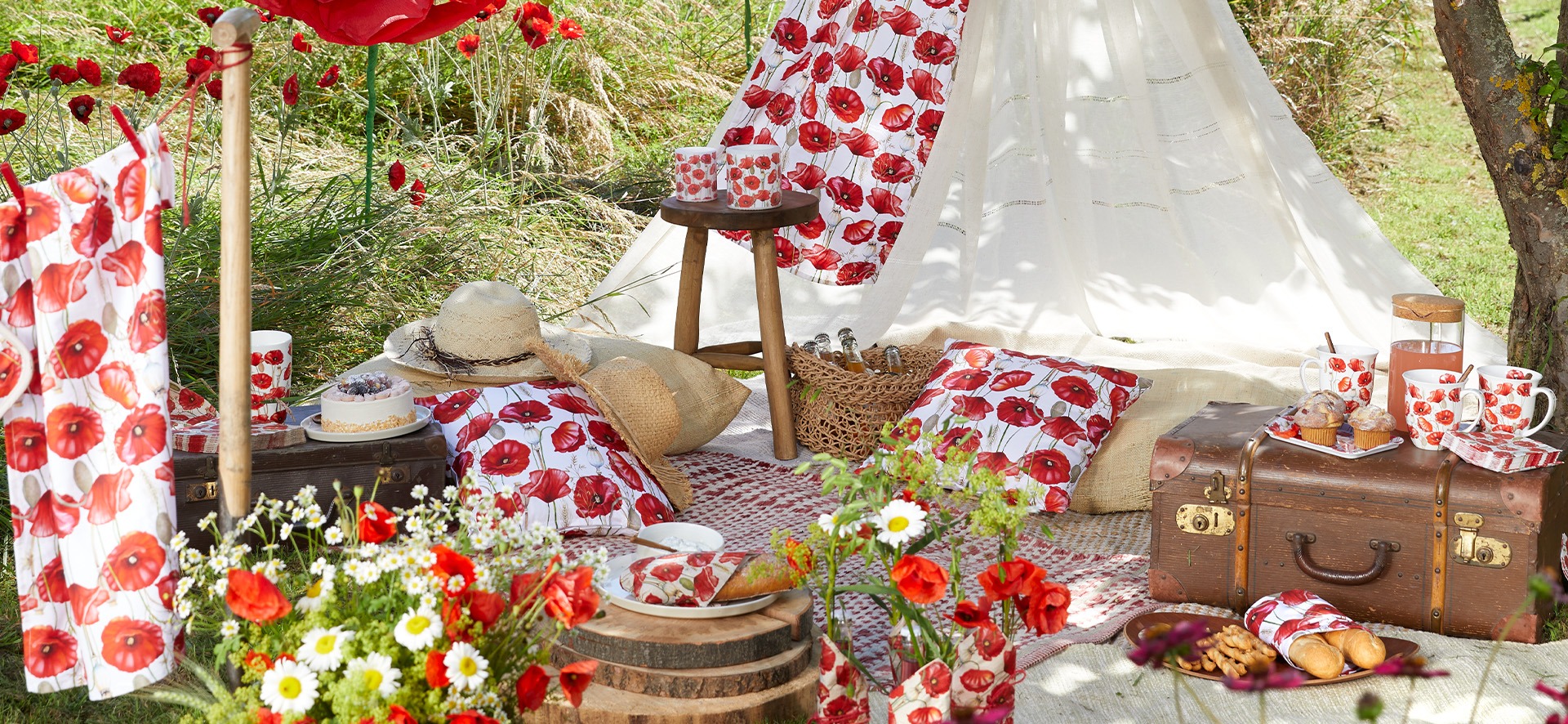 Close-up of a detailed garden picnic setup on grass, featuring a vintage suitcase, teepee, and various items adorned with red poppy patterns from the 'Poppy all over' series.