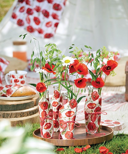 Glass bottles with flowers wrapped in 'Poppy all over' napkins on a wooden tray.